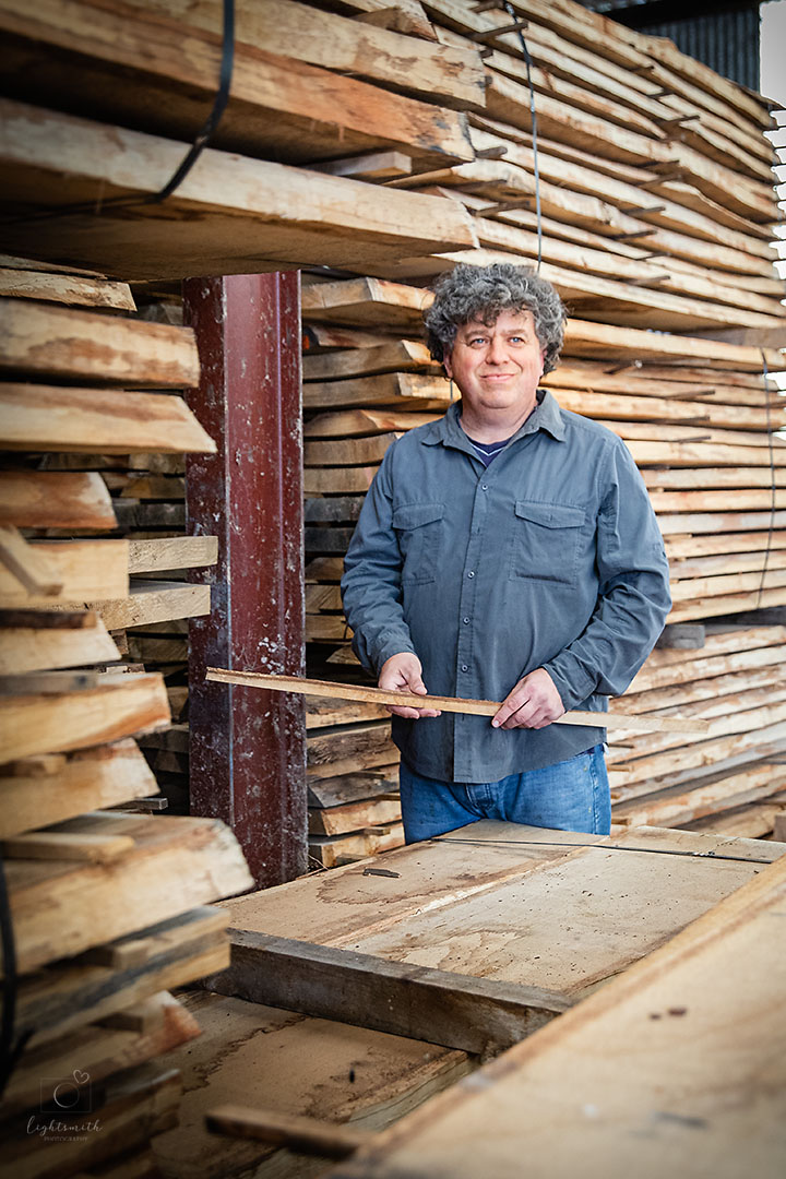an environmental portrait of a man in a wood depot. the wood is stacked high behind him, he is smiling