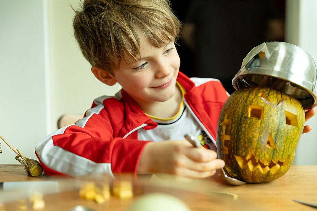 a boy smiling at the pumpkin he has carved. The pumpkin is wearing a metal bowl as an army helmet. 
