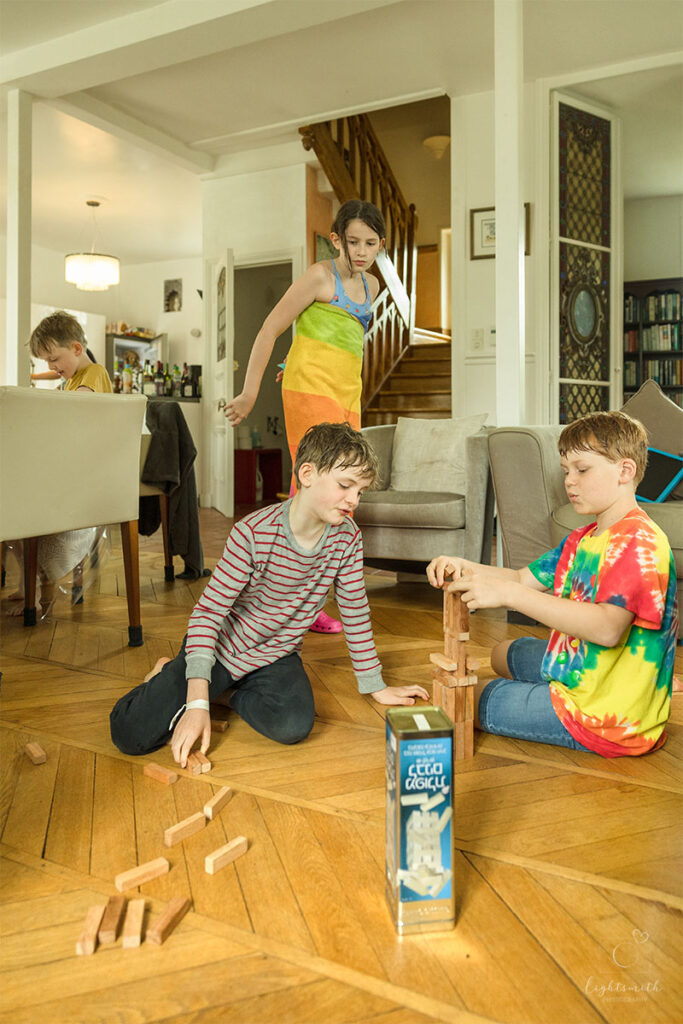 two boys playing jenga, a girl is walking by looking at their game. Another small boy is in the background drawing at the table. 