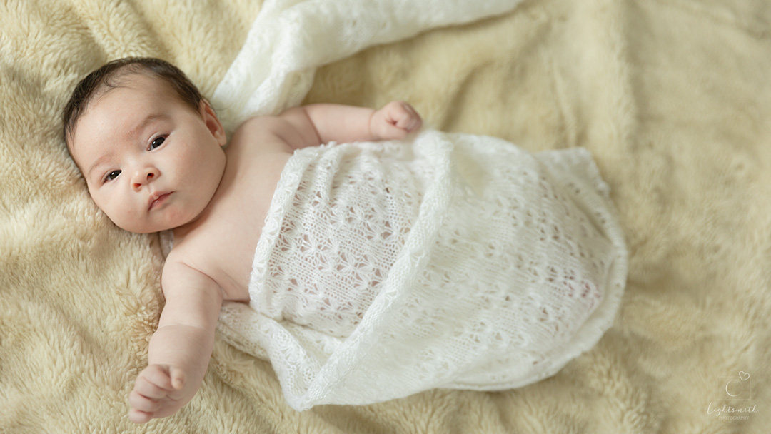 a newborn baby wrapped up in a cream lace blanket. The baby is looking at the camera and doing a superman pose!